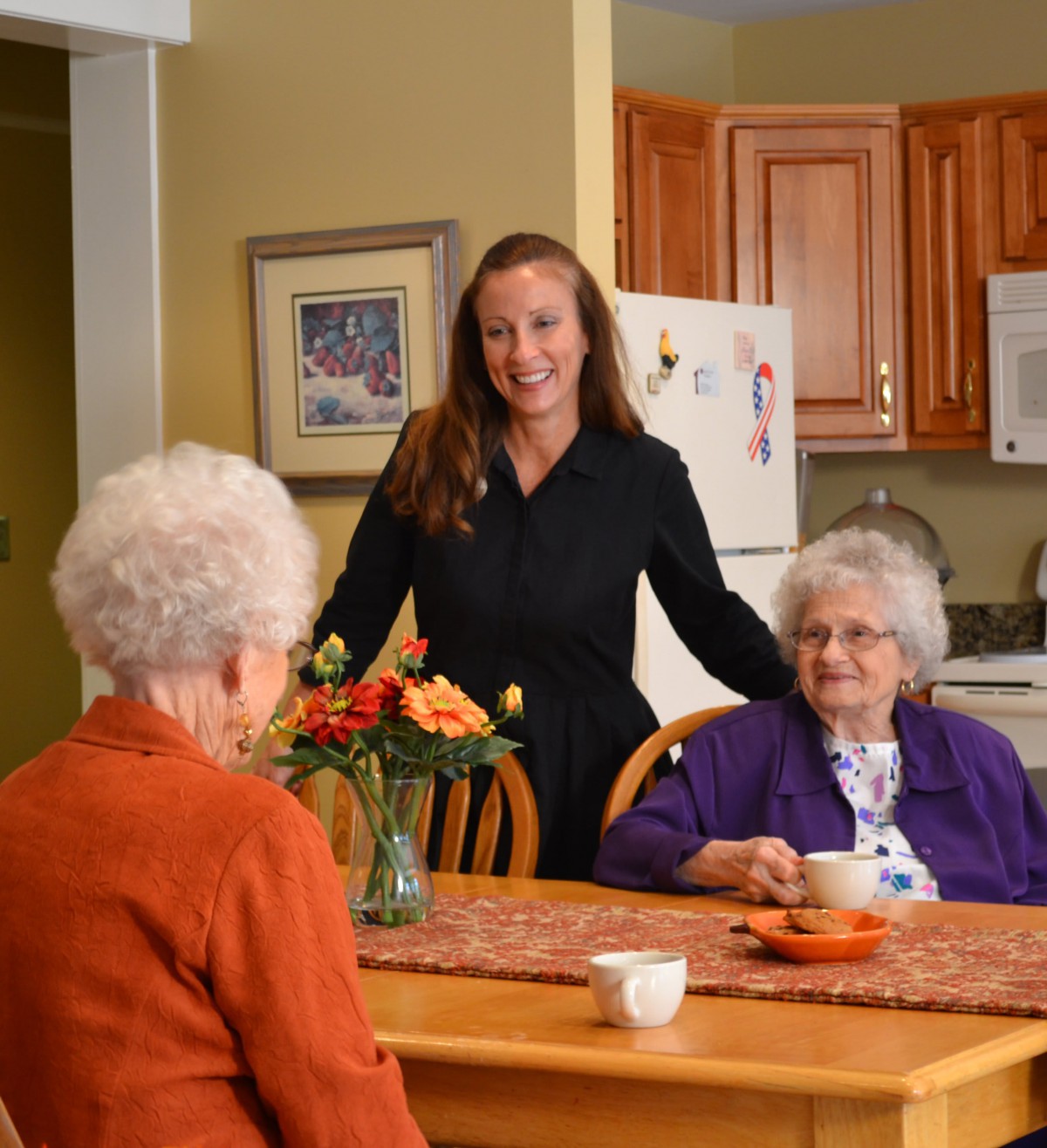 A caregiver interacting with two elderly women sitting at a kitchen table.