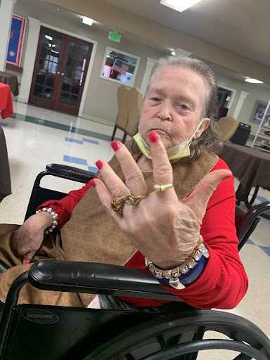 An elderly woman showcases her stylish red nails and accessories while sitting in a wheelchair.