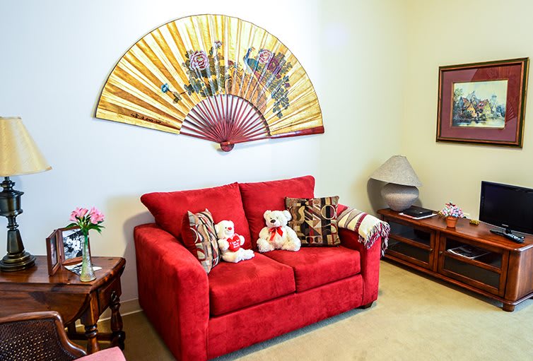 A cozy living room with a vibrant red sofa adorned with decorative cushions and teddy bears, set against a backdrop of an ornate wall fan.