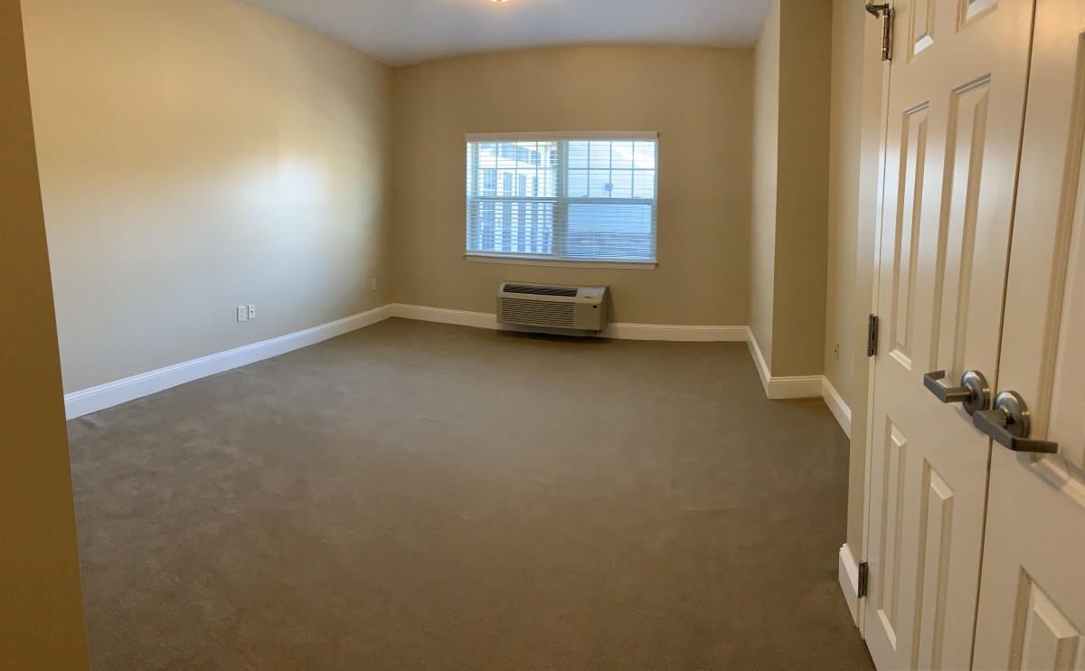 An empty room with beige walls, a carpeted floor, and a window with an air conditioning unit below it.