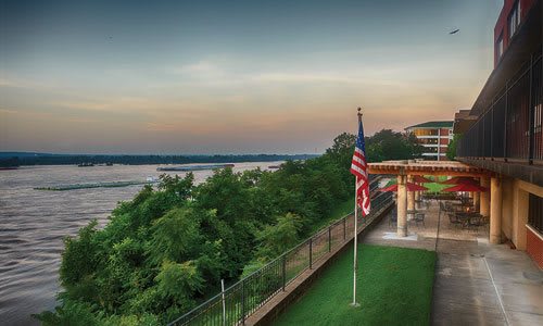 A scenic view of a river at sunset with an American flag and a building overlooking lush greenery.