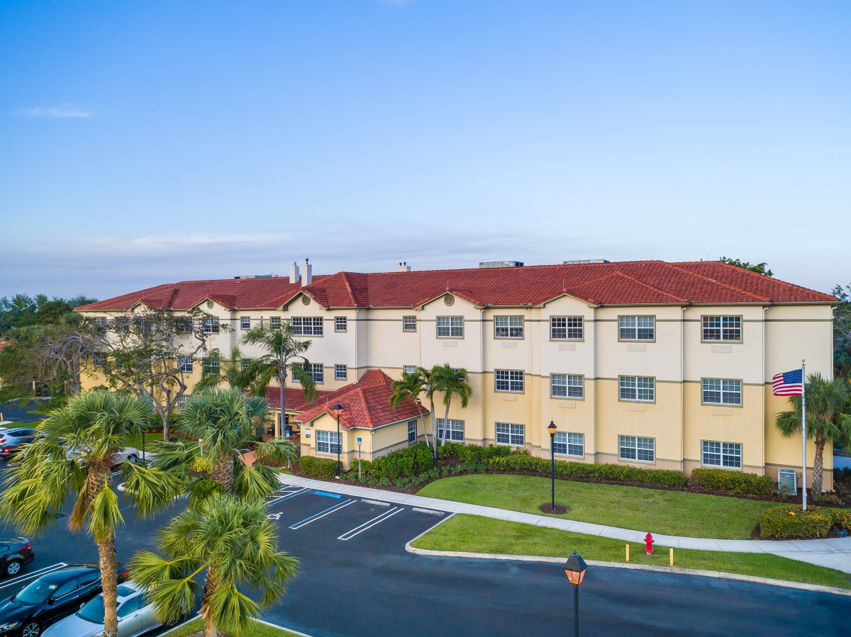 A residential apartment building with palm trees and a parking lot in the foreground.