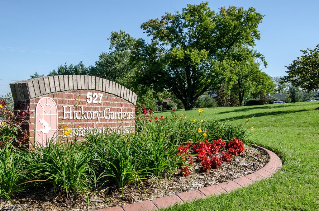 A brick sign with landscaping features vibrant flowers and lush greenery in a park setting.