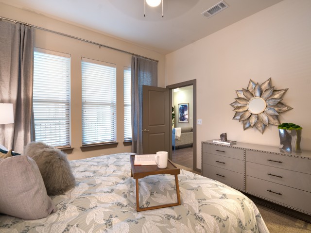 A modern bedroom with a neatly made bed featuring a decorative tray, a dresser with a stylish mirror, and large windows with blinds.