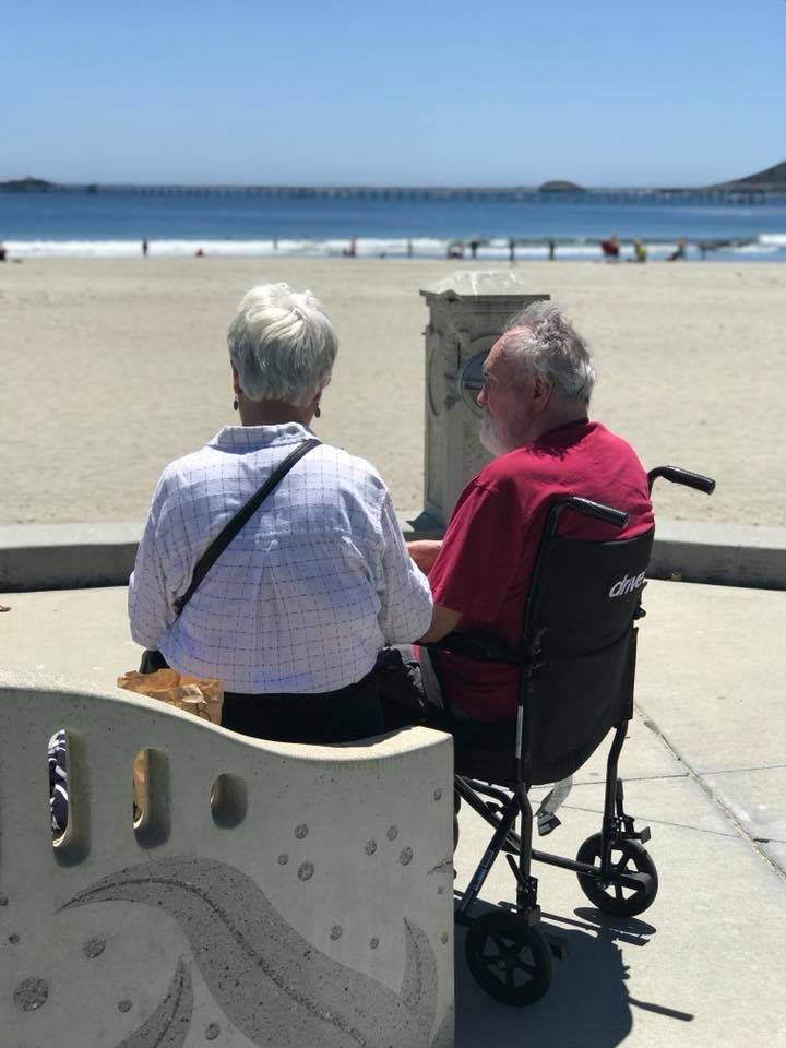 An elderly couple enjoys a peaceful day at the beach, with one seated in a wheelchair.