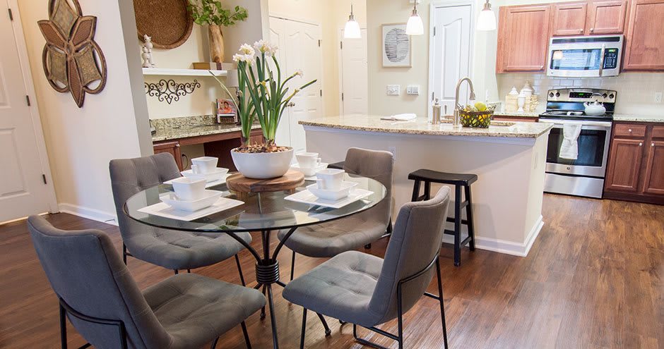 A cozy dining area with a round glass table set for four, featuring a centerpiece plant and modern gray chairs adjacent to a kitchen with bar stools.