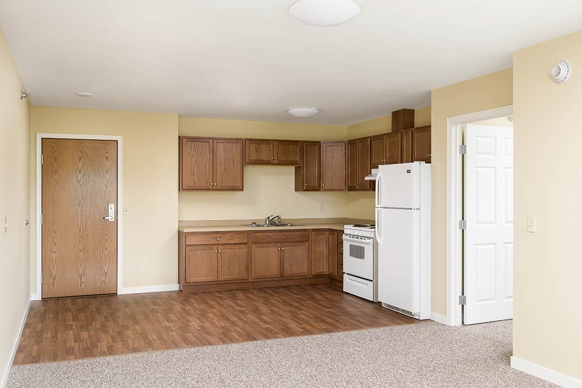 A small, empty apartment kitchen with wooden cabinets and basic appliances.