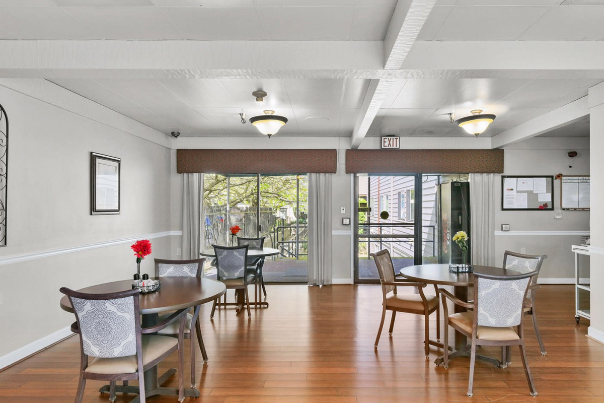 A spacious dining area with wooden floors, several tables and chairs, chandeliers, and large windows with curtains.