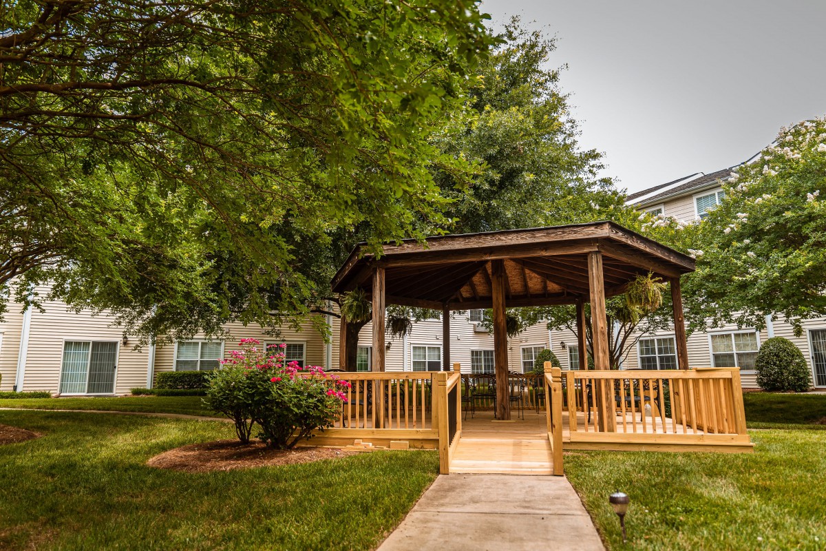 A wooden gazebo surrounded by lush greenery and a paved pathway in a garden setting.