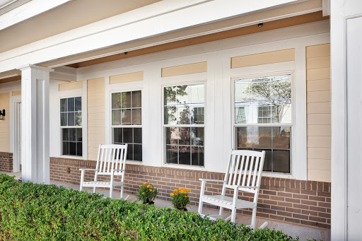 A cozy porch featuring two white rocking chairs beside vibrant plant pots and large windows.