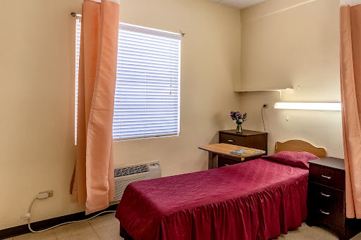 A simple, well-lit room featuring a single bed with a maroon bedspread, a desk, and natural light coming through the window.