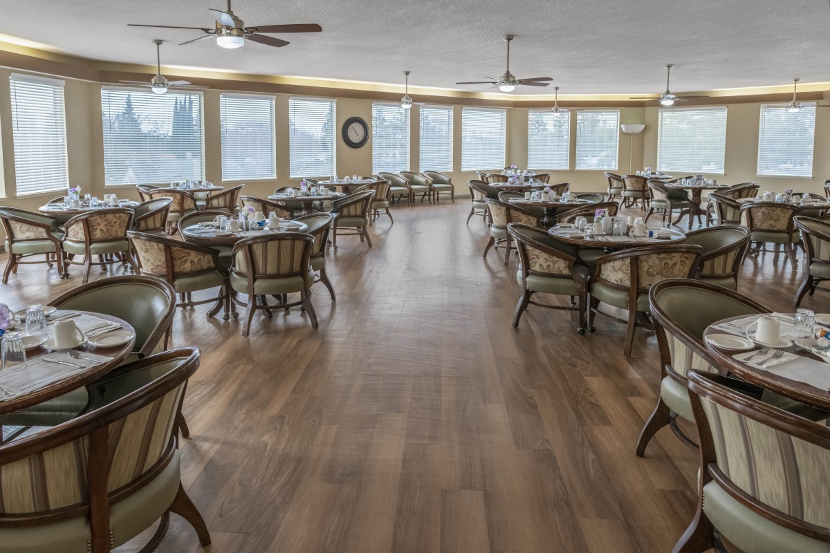 A spacious dining room with multiple tables and chairs, featuring large windows and ceiling fans.