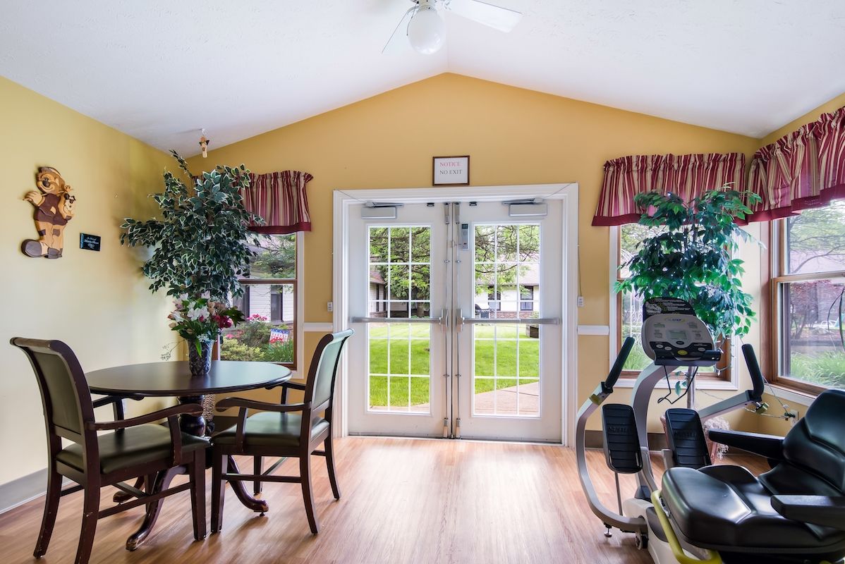 A bright and airy room with large windows, featuring a table with chairs, potted plants, and exercise equipment.