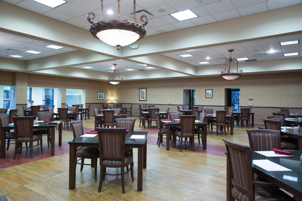 An empty dining hall with wooden tables and chairs, illuminated by chandeliers.