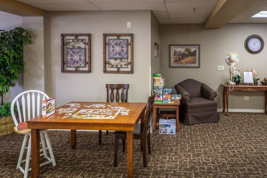 A cozy indoor space featuring a puzzle on a wooden table, surrounded by chairs and an armchair, with artwork adorning the walls.