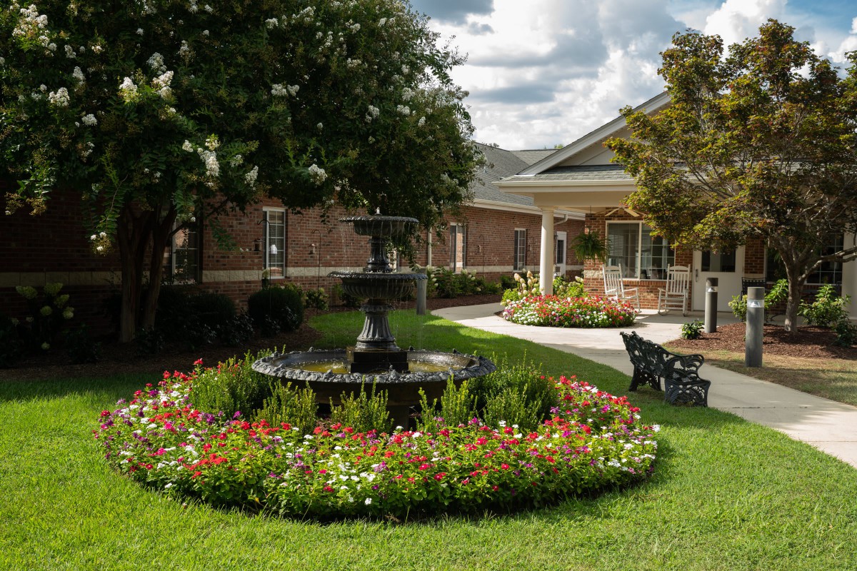 A serene garden with a central fountain, surrounded by colorful flowers and greenery, next to a building.