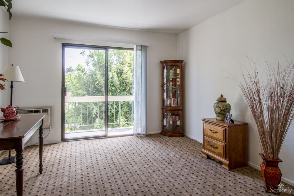 A bright living room with a sliding glass door leading to a balcony, featuring carpeted flooring and wooden furniture.