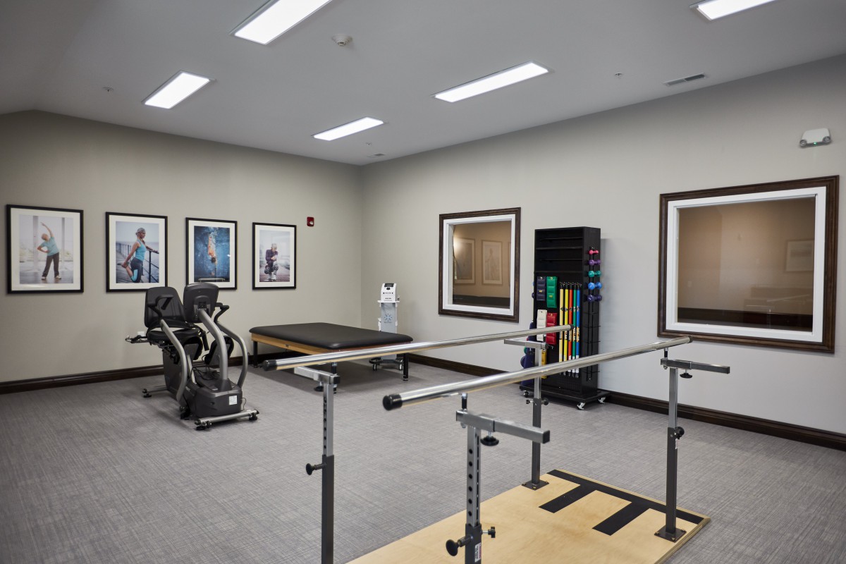 A rehabilitation room equipped with exercise machines, a therapy bed, parallel bars, and mirrors.