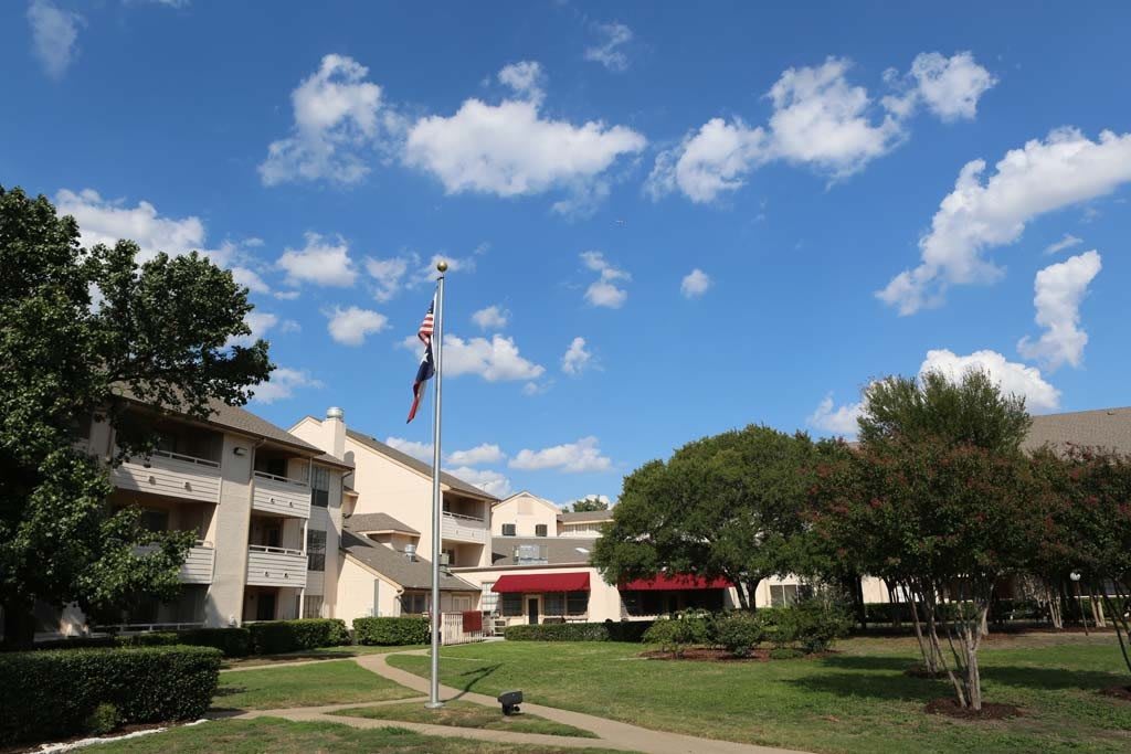 A scenic view of a building complex with a flagpole, surrounded by trees and under a blue sky with fluffy clouds.