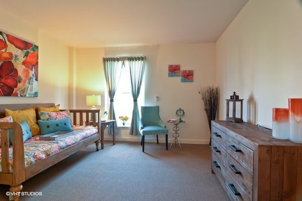 A cozy bedroom with a comfortable armchair, a window with curtains, and a wooden dresser.