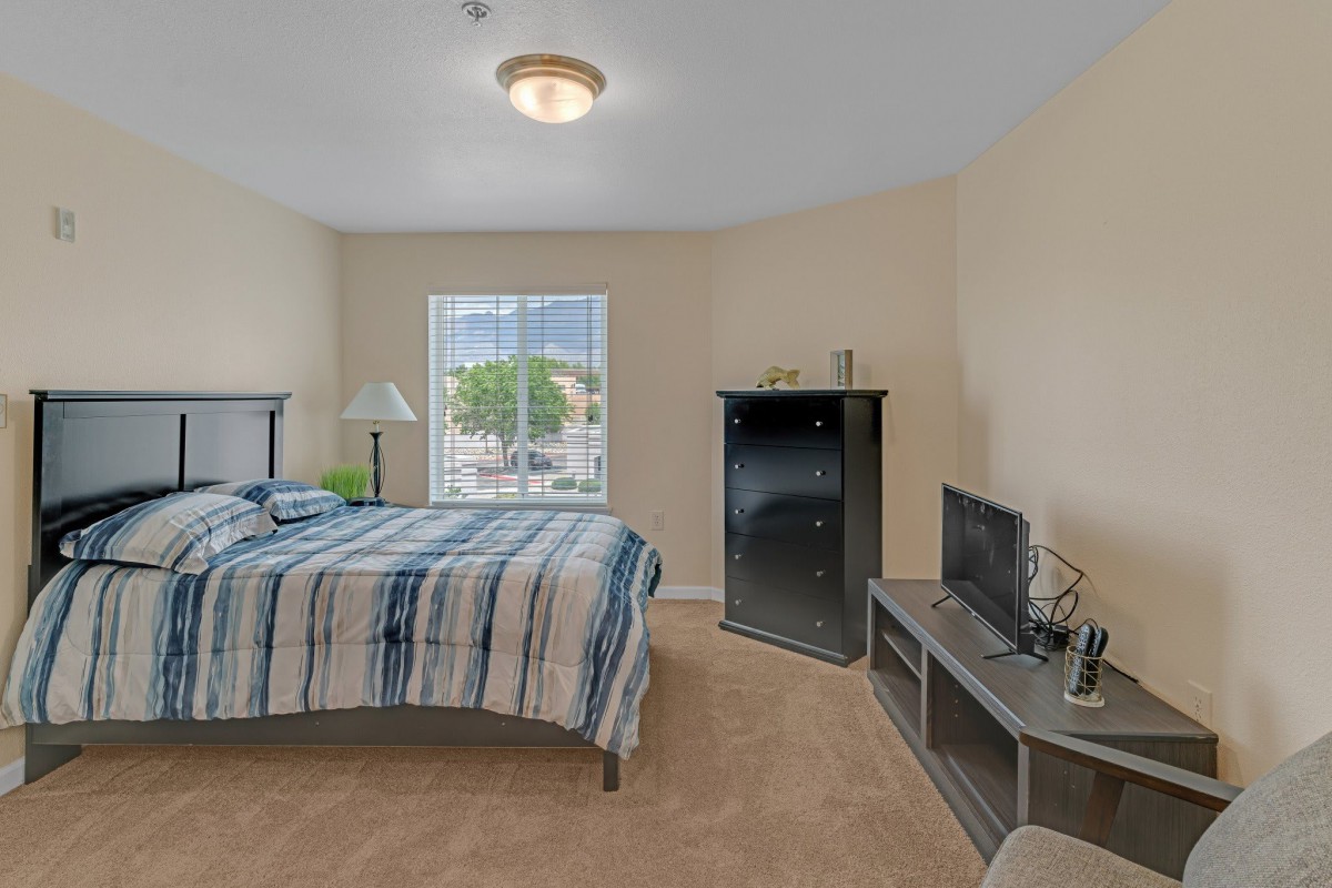 A cozy bedroom with a striped bedspread, featuring a window, lamp, TV stand, and carpeted floor.