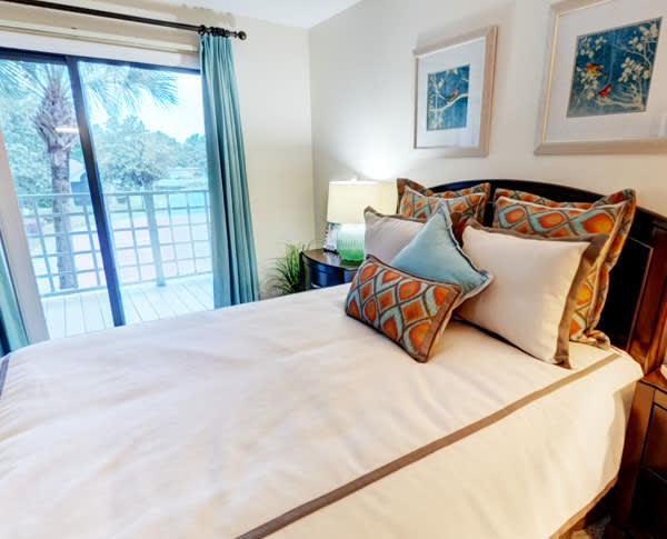 A cozy bedroom with a neatly made bed and colorful pillows, featuring a window with curtains and decorative artwork on the walls.