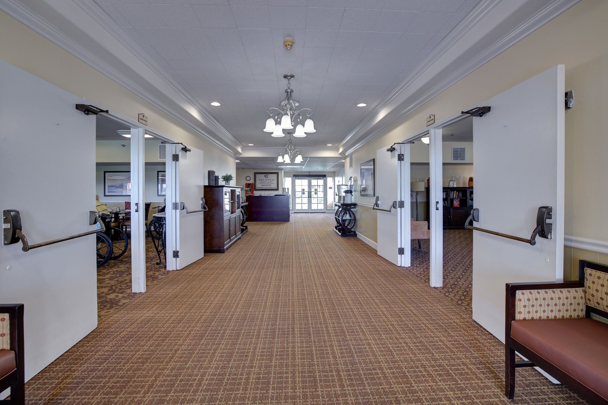 A spacious, carpeted hallway with chandeliers and open doors, illuminated by natural light.