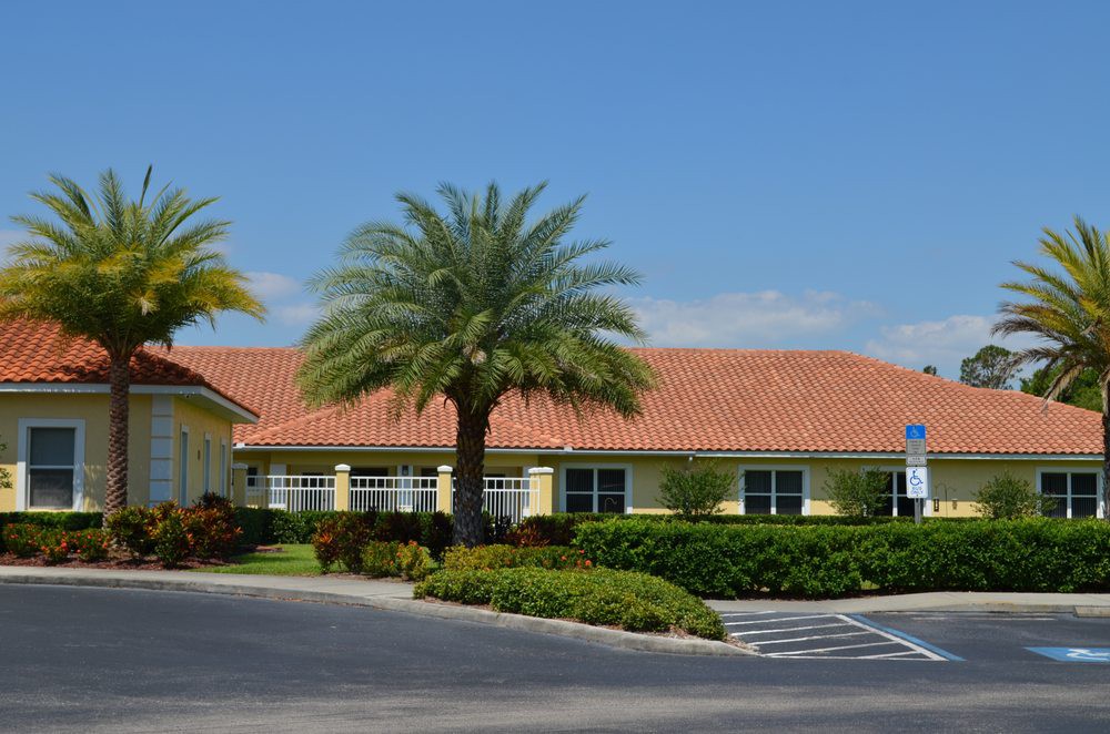 A building with a tile roof, surrounded by palm trees and landscaping, adjacent to a parking lot under a clear blue sky.