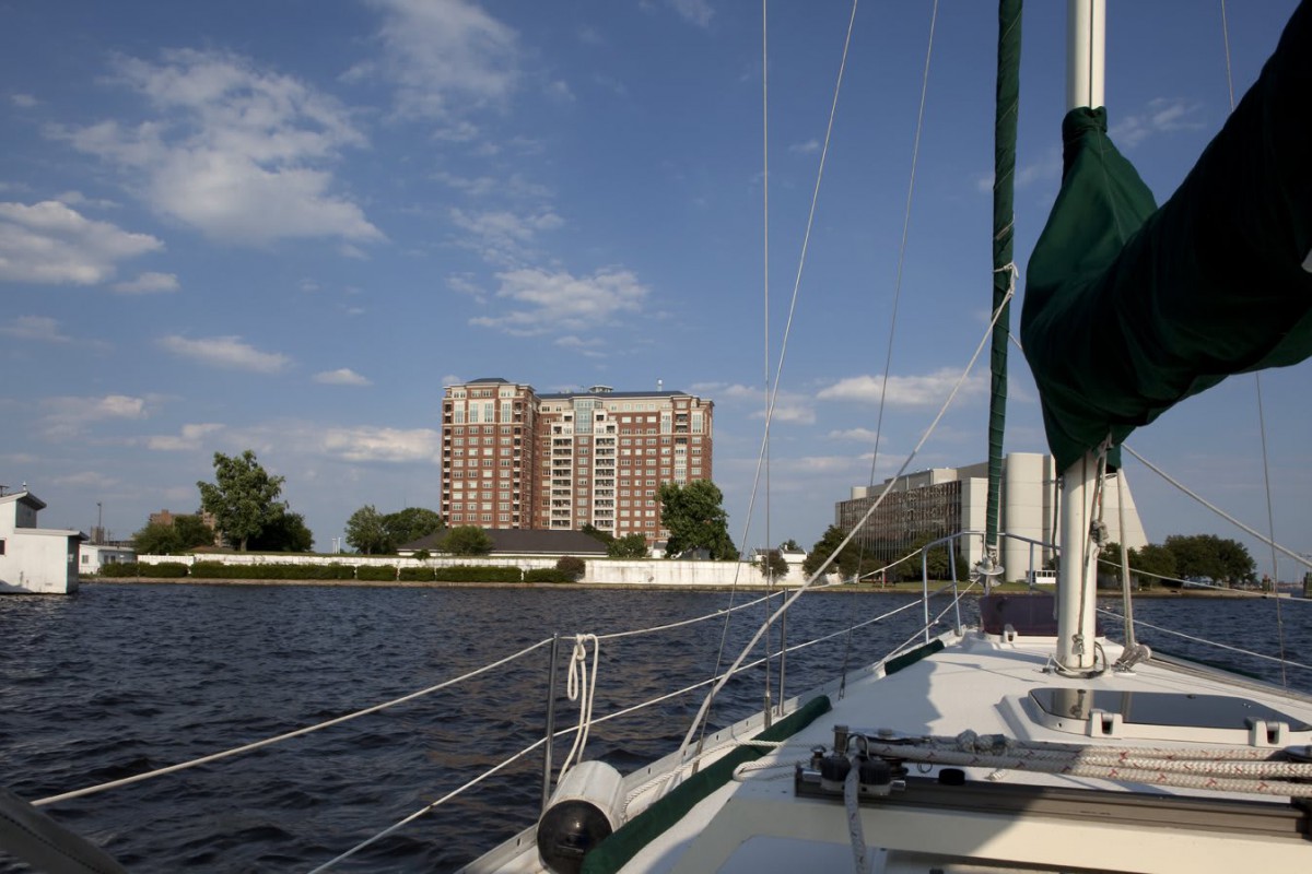 A sailboat cruising on a river with a city building in the background under a partly cloudy sky.