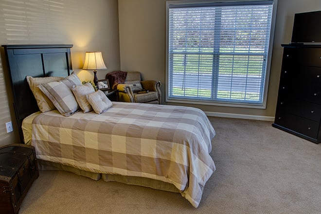 A cozy bedroom with a neatly made bed, a large window, and a comfortable chair.