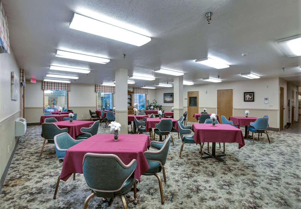 A spacious dining area with multiple tables covered in red tablecloths and surrounded by chairs, illuminated by fluorescent lighting.