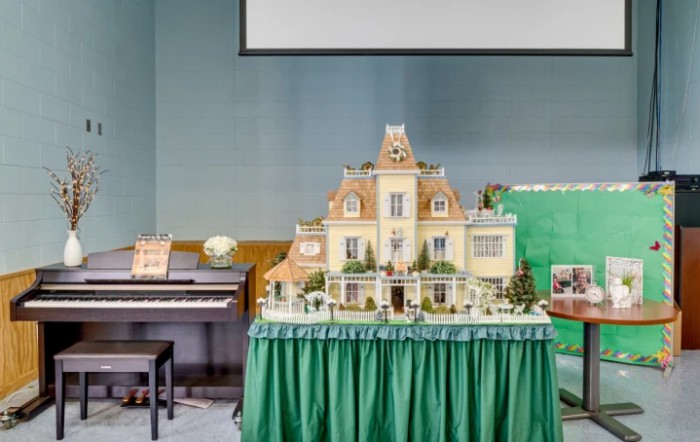 A detailed dollhouse model displayed on a table with a green skirt, positioned next to a piano in an exhibition setting.