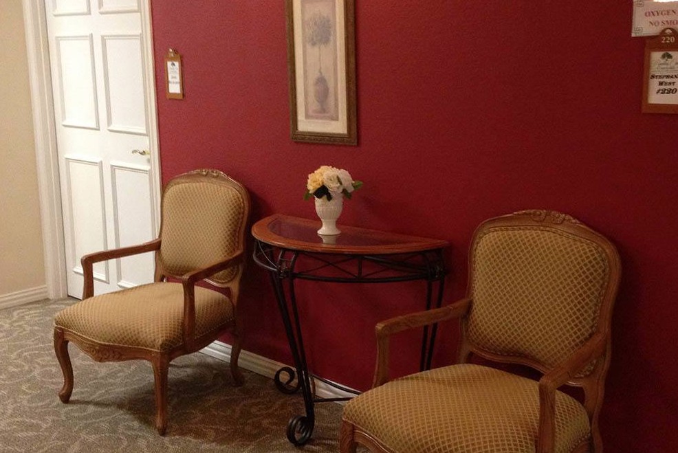 A cozy waiting area with two chairs, a table adorned with flowers, and a framed picture on a red wall.