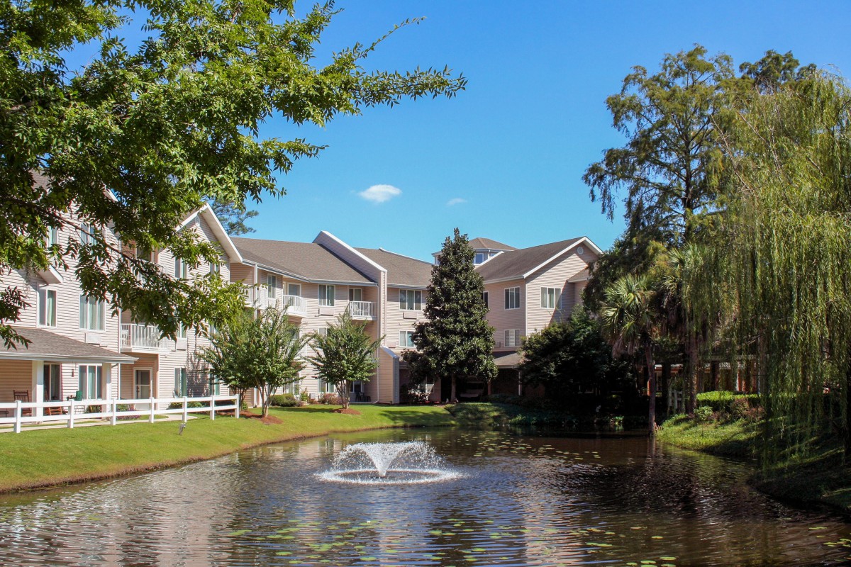 A serene residential building with a pond and fountain in the foreground, surrounded by trees under a clear blue sky.