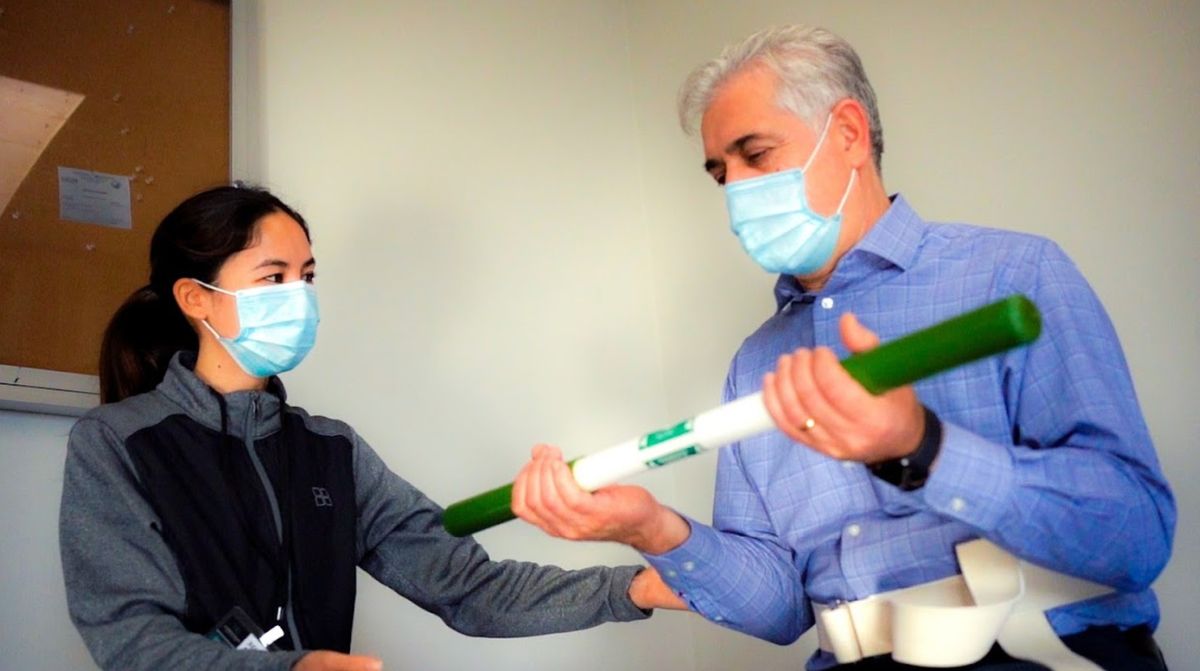 Two individuals wearing masks engage in an exercise or physical therapy session with a resistance bar.