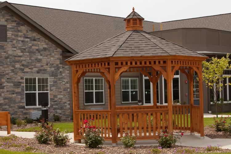 A wooden gazebo stands in front of a house, surrounded by landscaping.