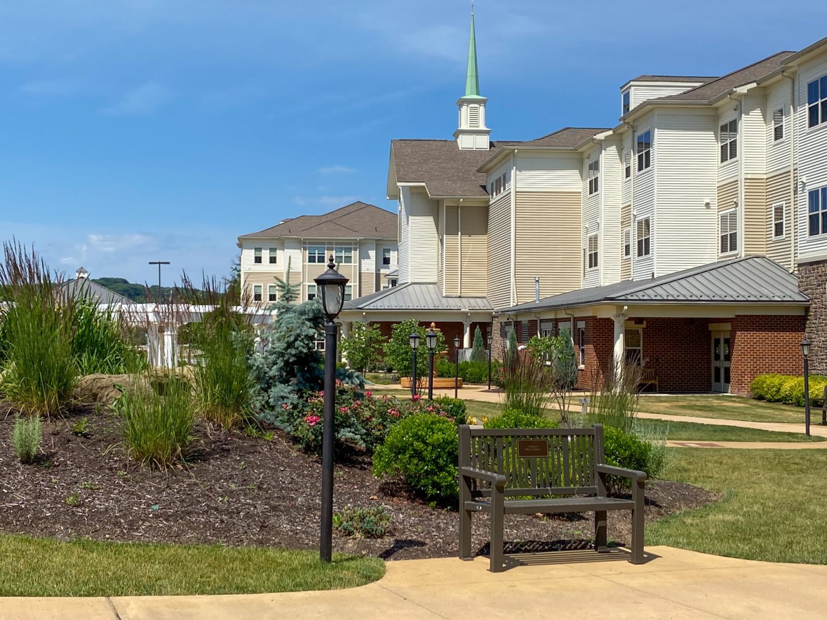 A well-maintained garden with a bench and pathway in front of a residential building with a church steeple in the background under a clear blue sky.