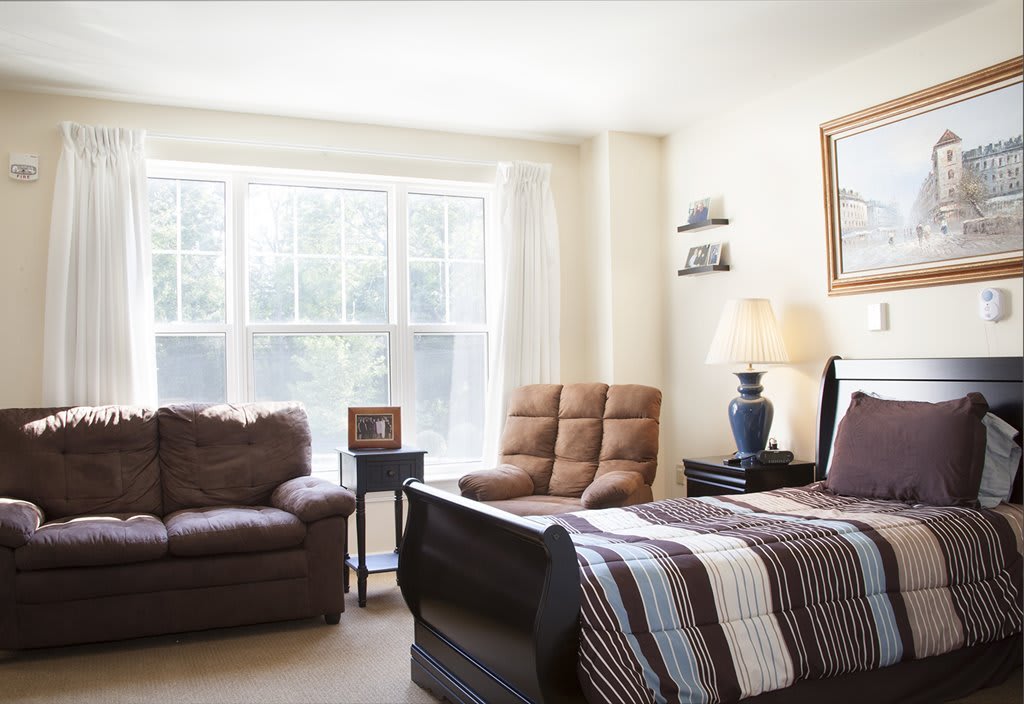 A cozy bedroom with a large window allowing plenty of natural light, featuring a bed, armchair, and sofa.