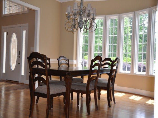 A cozy dining room with a wooden table and chairs, illuminated by a chandelier and natural light from large windows.