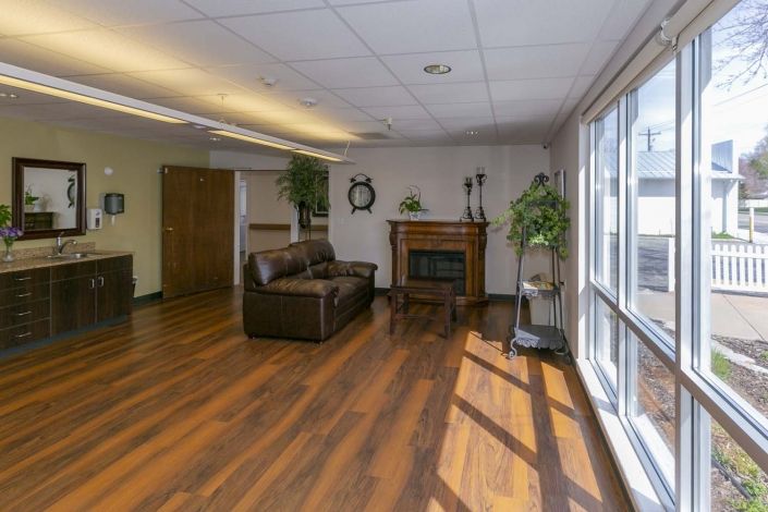 A spacious living room with hardwood flooring, a sofa set, and a fireplace, complemented by indoor plants and large windows letting in natural light.