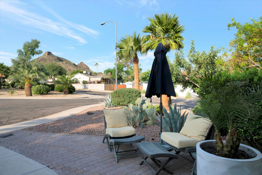 A patio with comfortable outdoor seating and an umbrella, surrounded by palm trees and desert plants in a residential neighborhood.