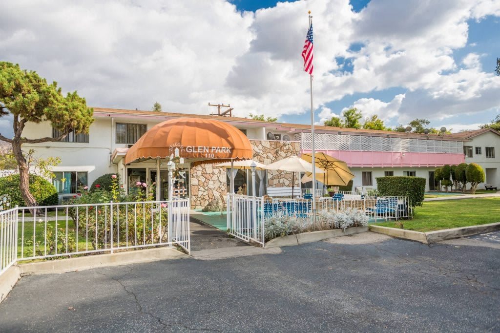 The exterior view of a roadside motel with an American flag, awning, and outdoor seating area under a partly cloudy sky.