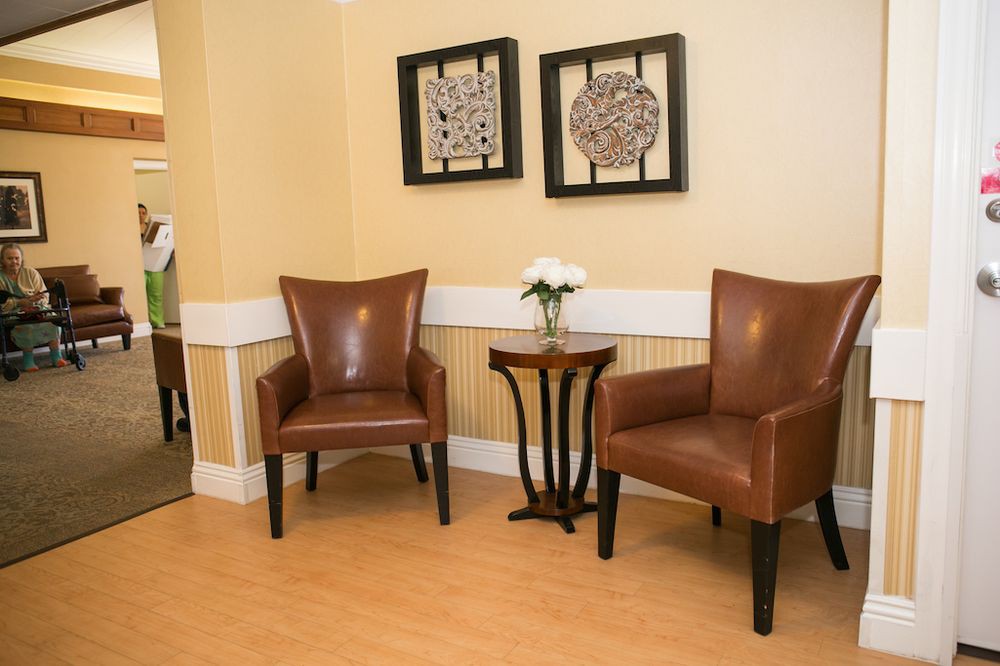 A cozy waiting area featuring two leather chairs, a small wooden table with a vase of flowers, and framed artwork on the wall.