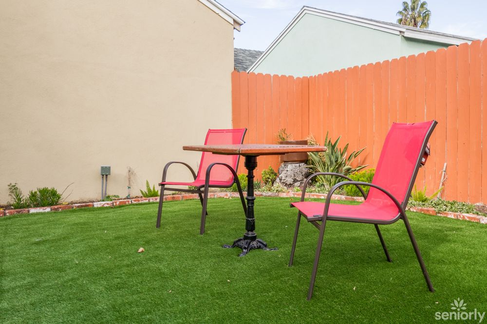 A cozy backyard setup with red chairs and a table on artificial grass, surrounded by a wooden fence and plants.