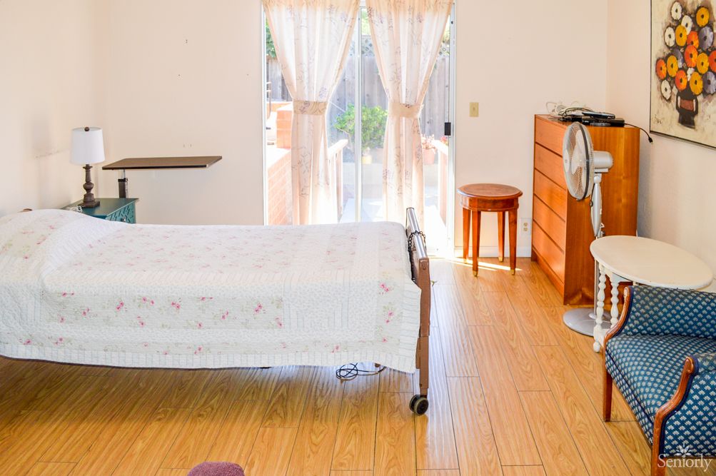 A well-lit bedroom with wooden flooring and simple furniture, including a bed, dresser, and stool.