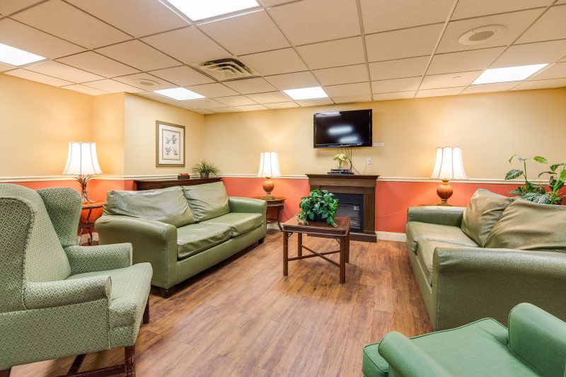 A cozy living room with green sofas, armchairs, a television mounted above a fireplace, and warm lighting.