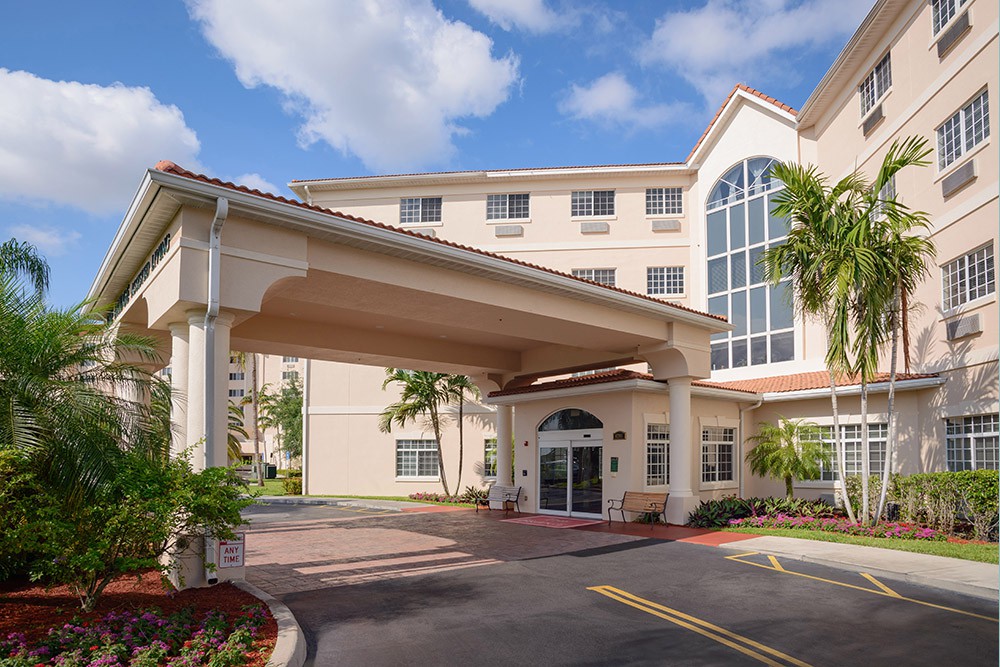 A modern hotel entrance with palm trees and a covered driveway under a partly cloudy sky.