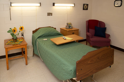 A simple patient room featuring a bed with green bedding, a bedside table, and a comfortable chair.