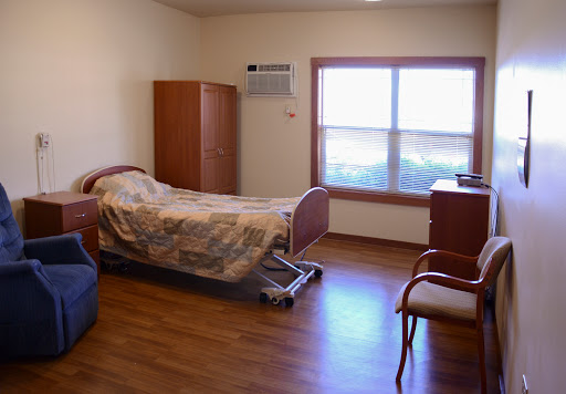 A clean and simple hospital room featuring a patient bed, a chair, and a window providing natural light.