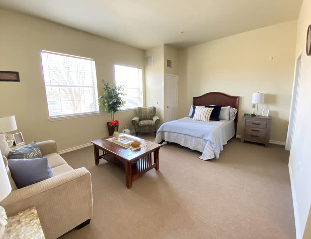 A well-lit bedroom with a bed, sofa, chairs, and a coffee table on a carpeted floor.